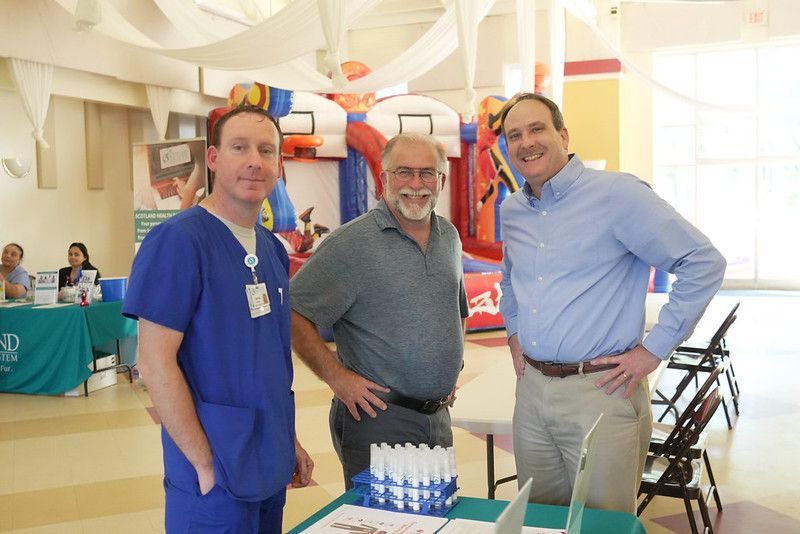 Three men smiling, standing behind a table at an event, bouncy castle in background.