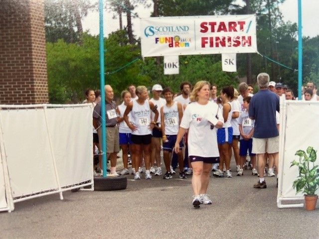 People at a 5K race start line, under a banner. Spectator walks in front.