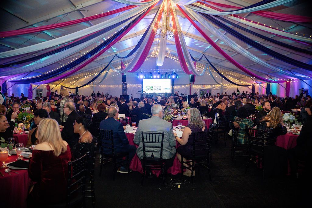 Large event tent filled with people at tables; red, white, and blue draped ceiling, stage in the back.