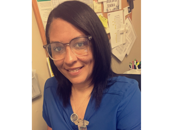 Woman with glasses wearing a blue top, smiling, in front of a bulletin board.