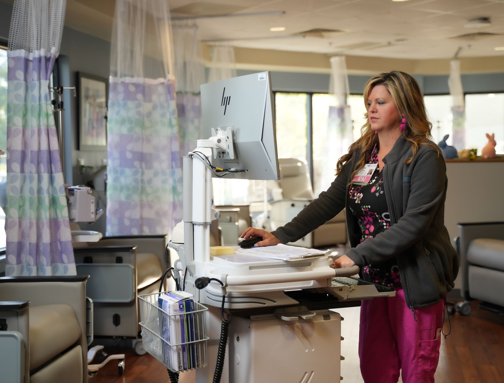 Nurse at a computer workstation in a patient care area, wearing pink scrubs and a gray jacket.
