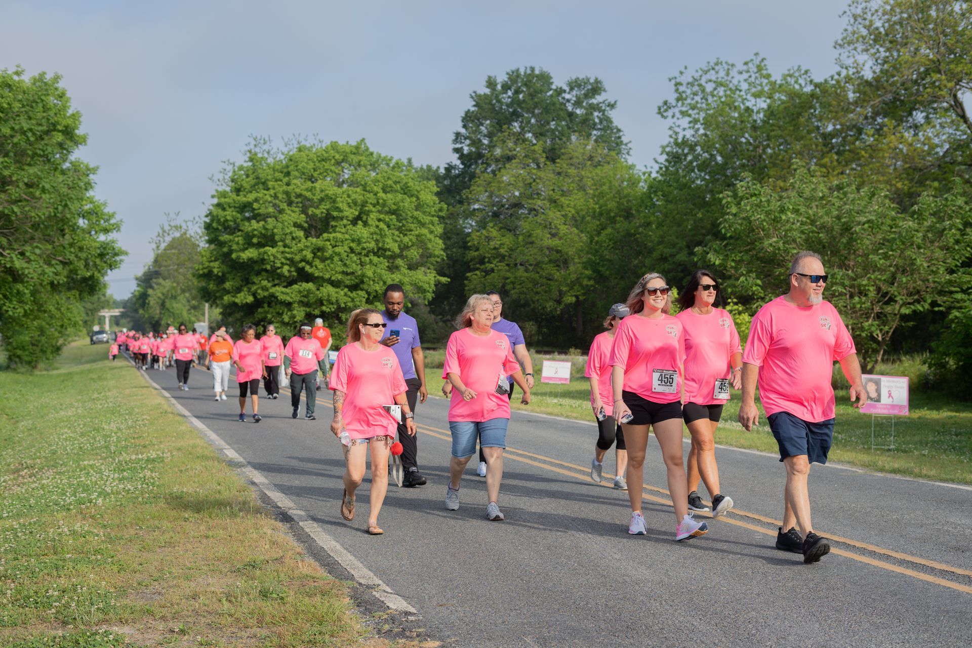 People in pink shirts walking on a road; likely a fundraising walk. Trees and greenery in the background.