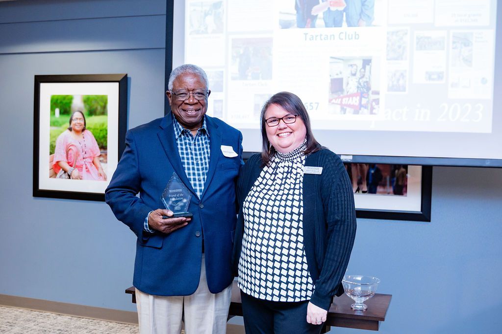An older man in a blue blazer and a woman in a black and white top stand together, smiling. They are holding an award.