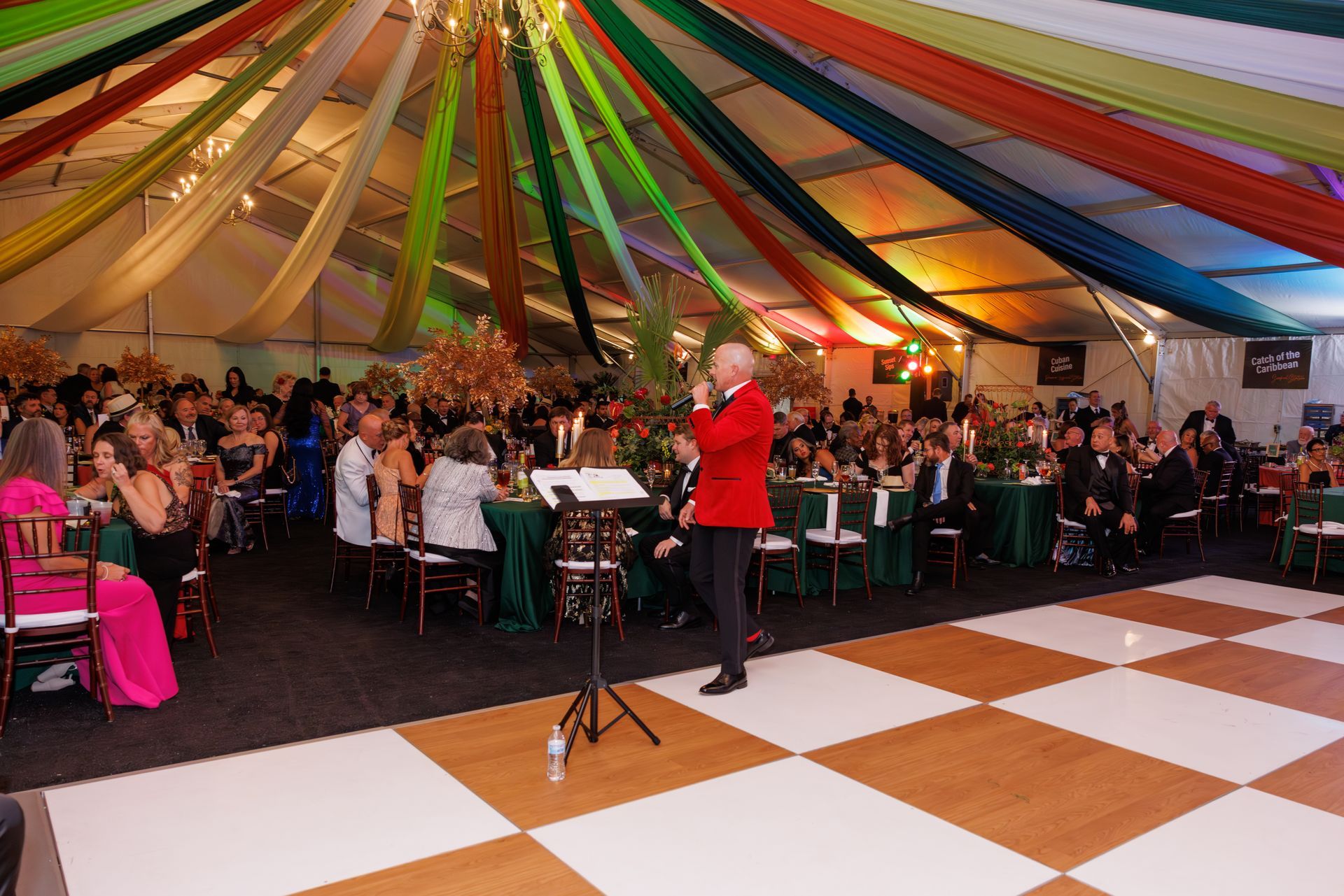 Man in red jacket sings at event, decorated tent with colorful fabrics, guests at tables, checkered dance floor.