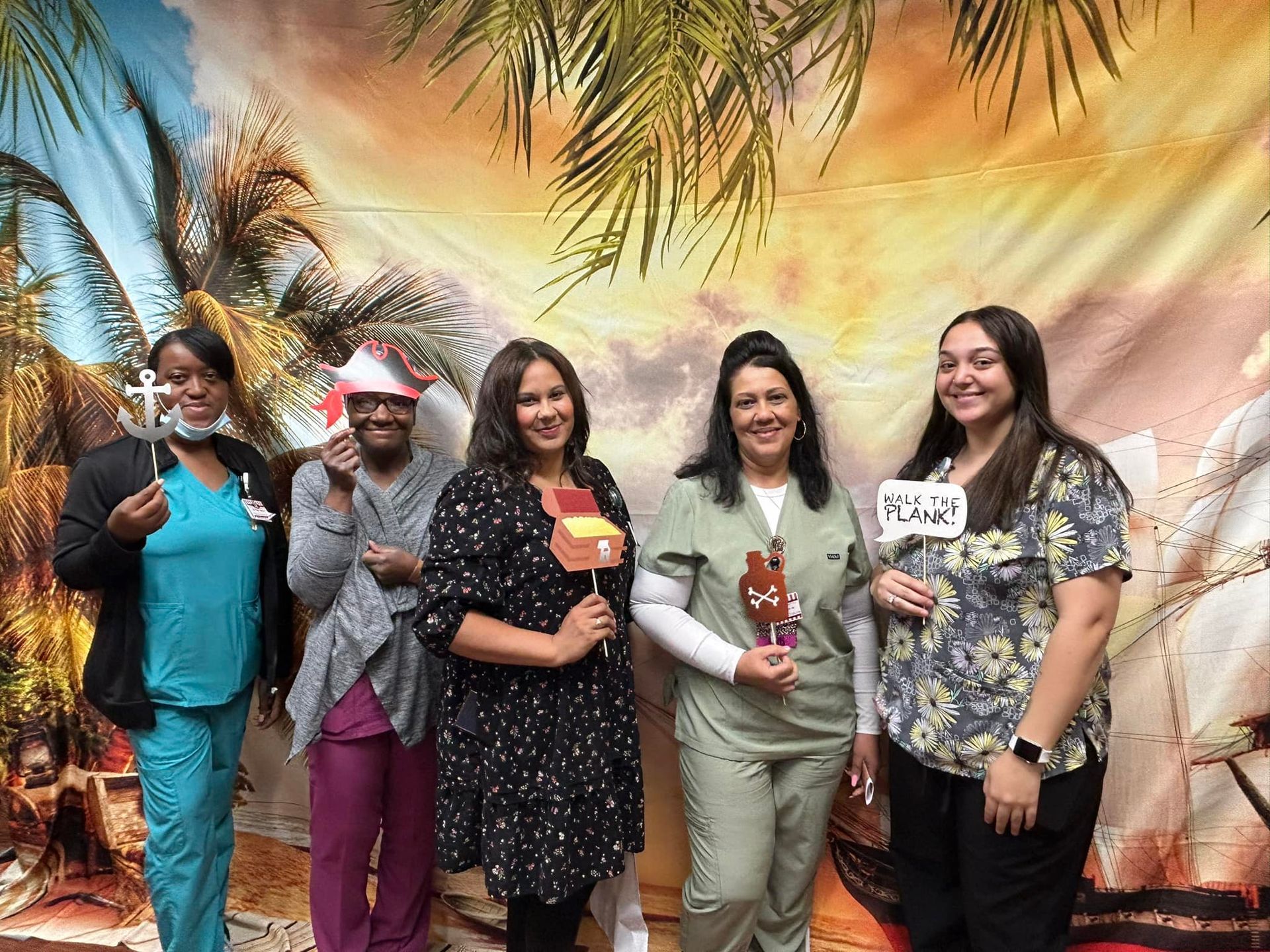 Five people posing in front of a tropical background. They are holding props like sunglasses, a treasure chest, and a sign.