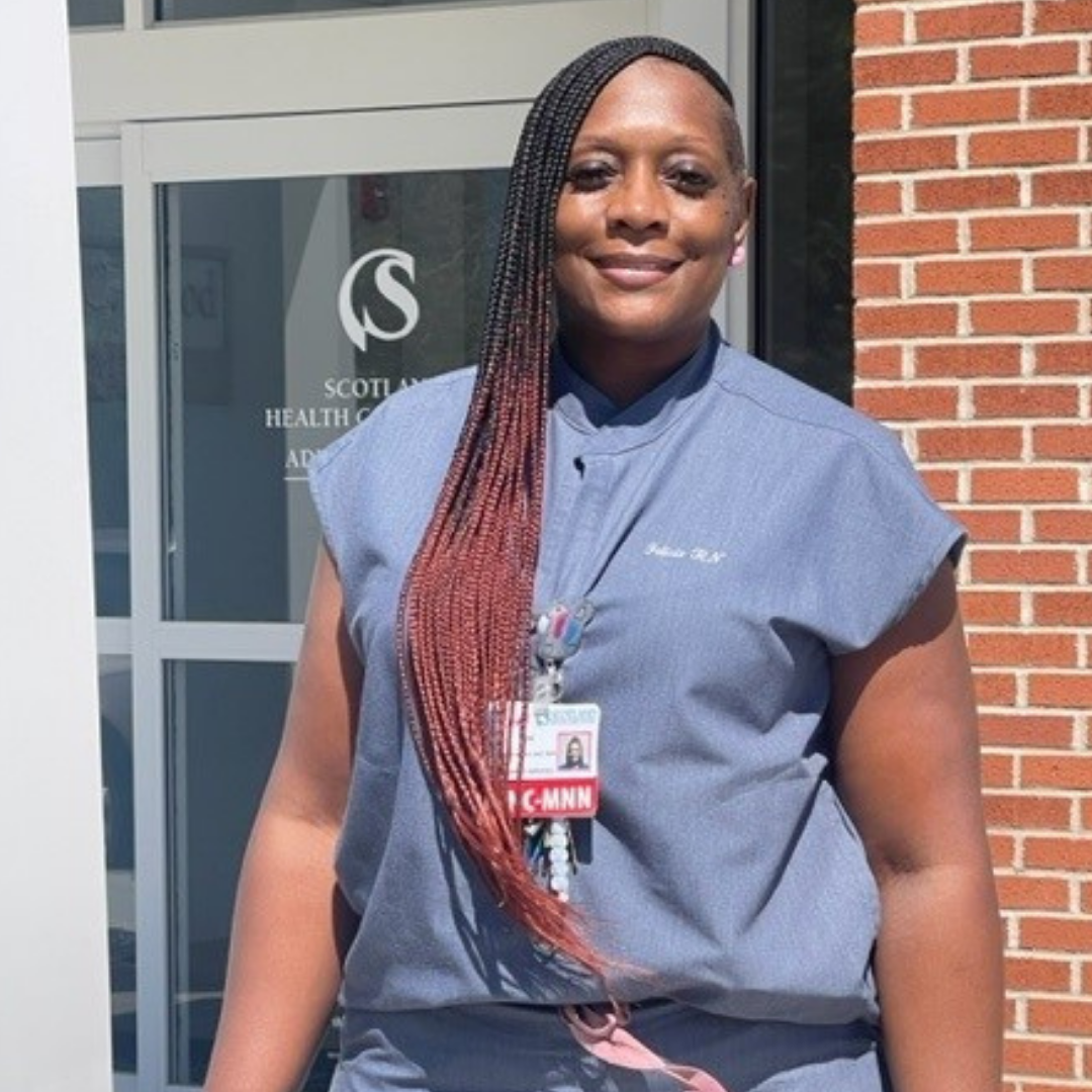Woman in medical scrubs stands outside a building. She has long braids with red tips and wears an ID badge.