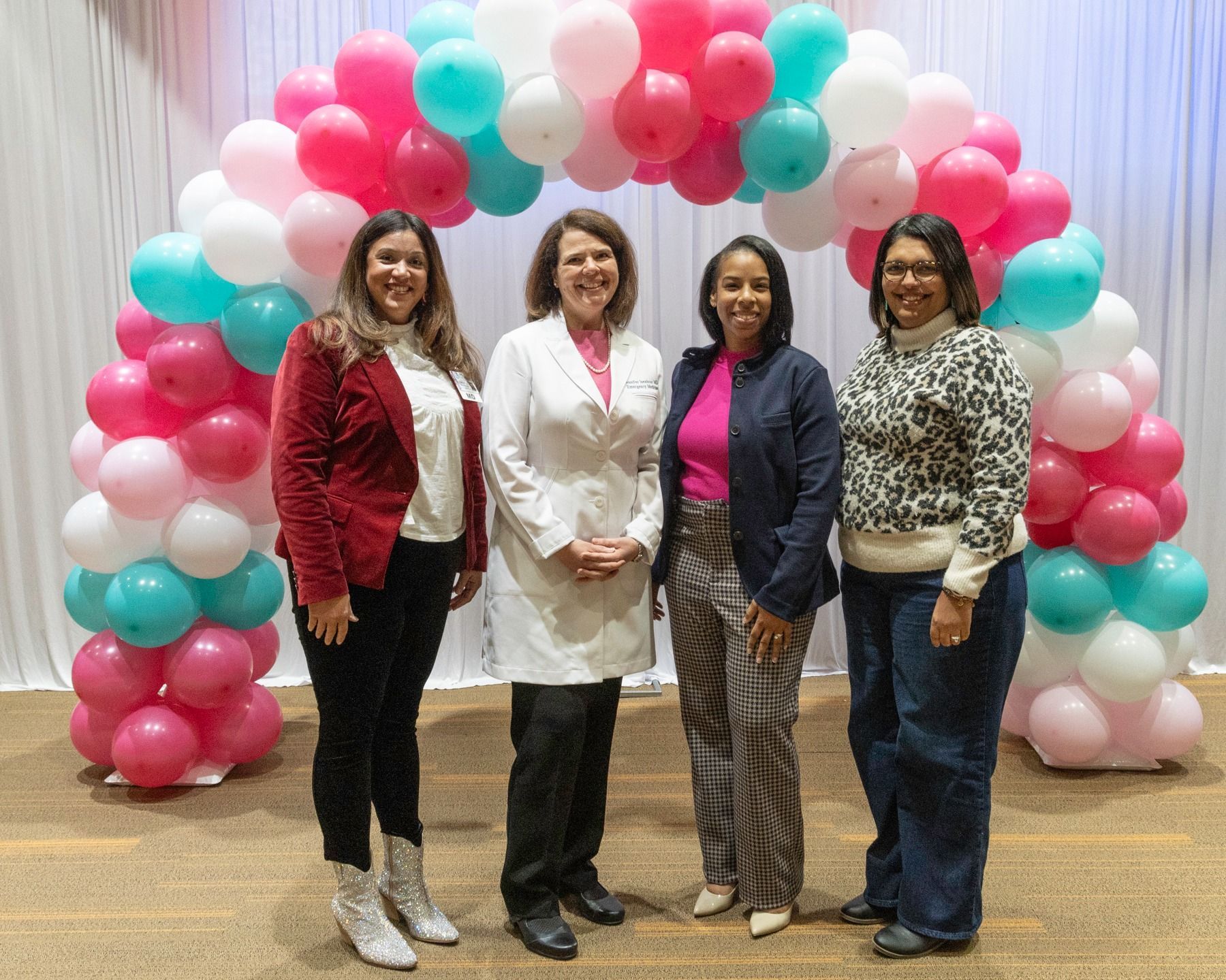 Four people stand smiling in front of a balloon arch with pink, white, and teal balloons.