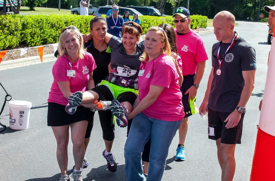 People in pink shirts carrying a person in athletic wear, likely an injured runner, at an outdoor event.