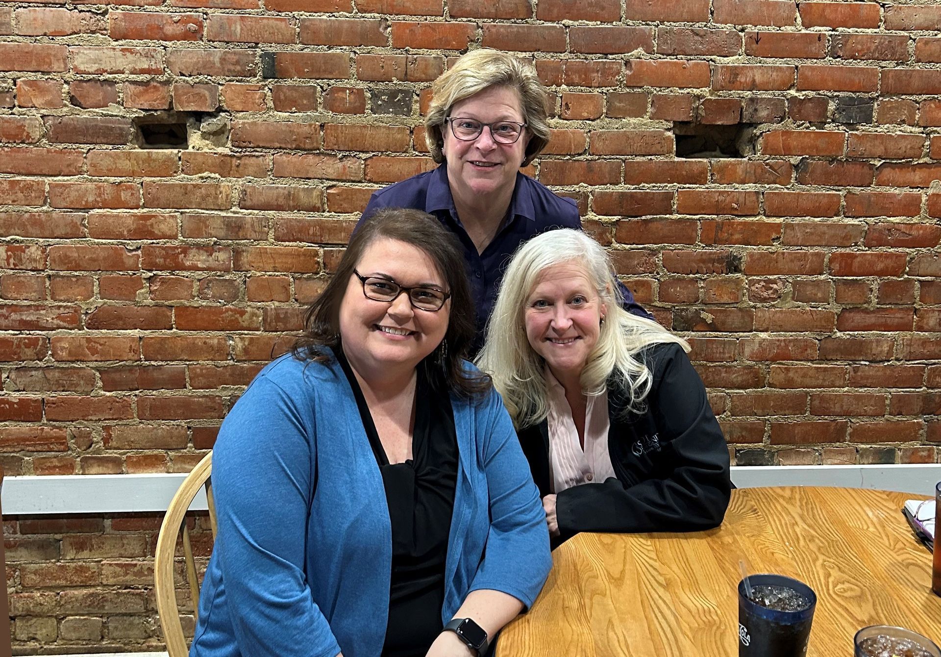 Three women seated at a table against a brick wall. Two are smiling at the camera.