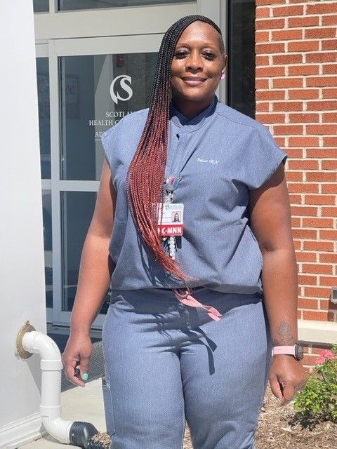 Person in blue uniform stands in front of a building with a logo. Braided hair has red highlights.