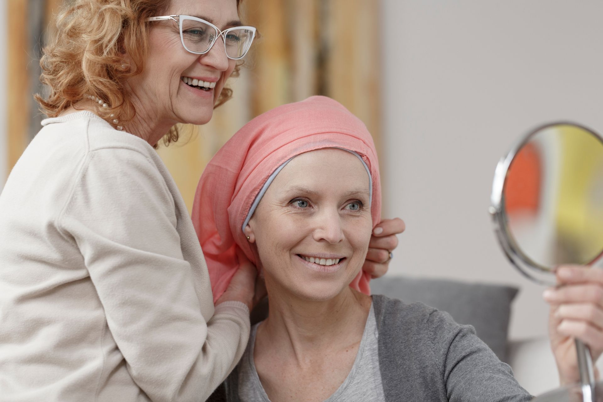 Woman with headscarf smiles at herself in mirror, assisted by another woman adjusting the scarf.