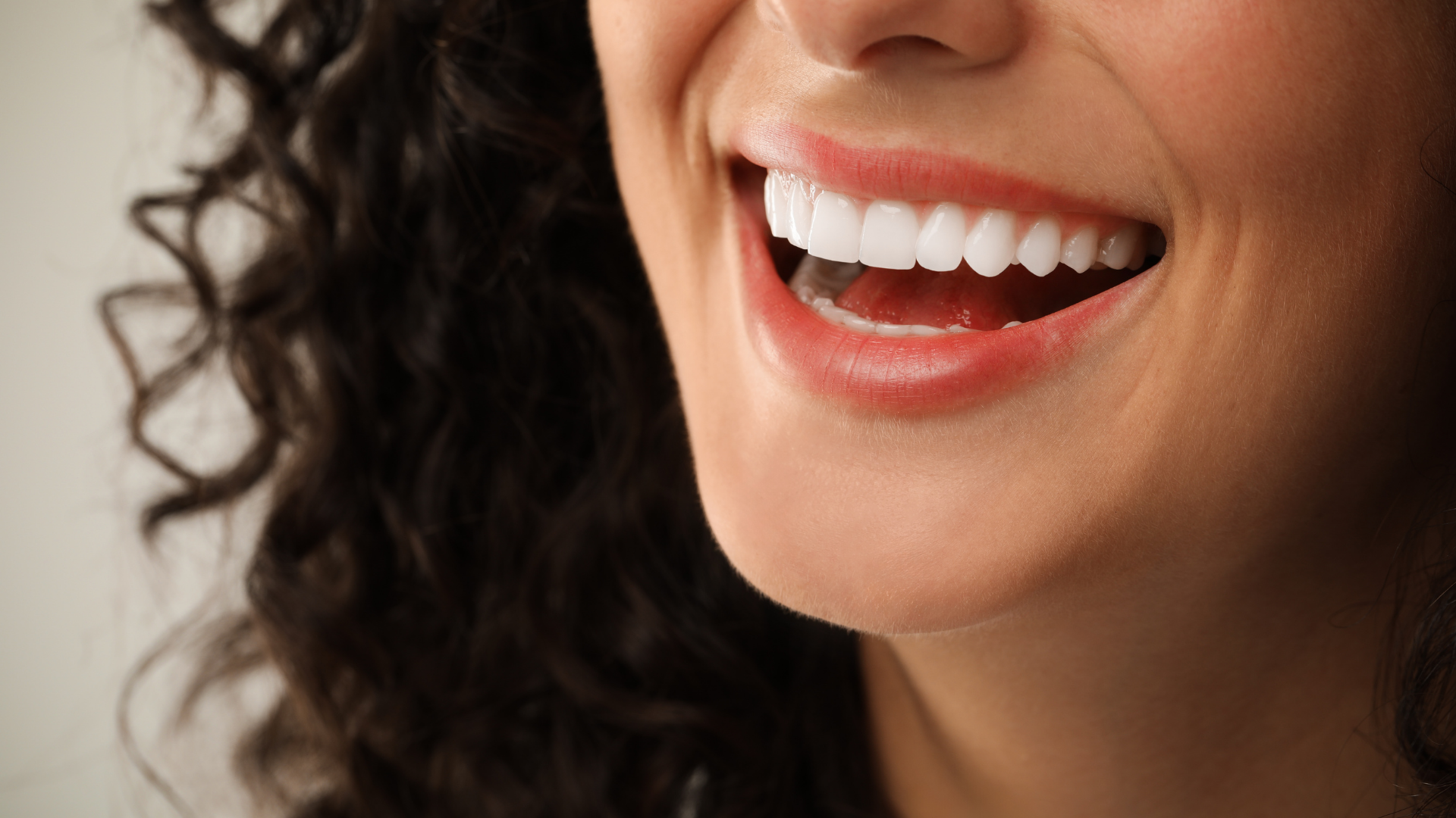 Close-up of a person smiling, showing white teeth, and pink gums. They have curly dark hair.