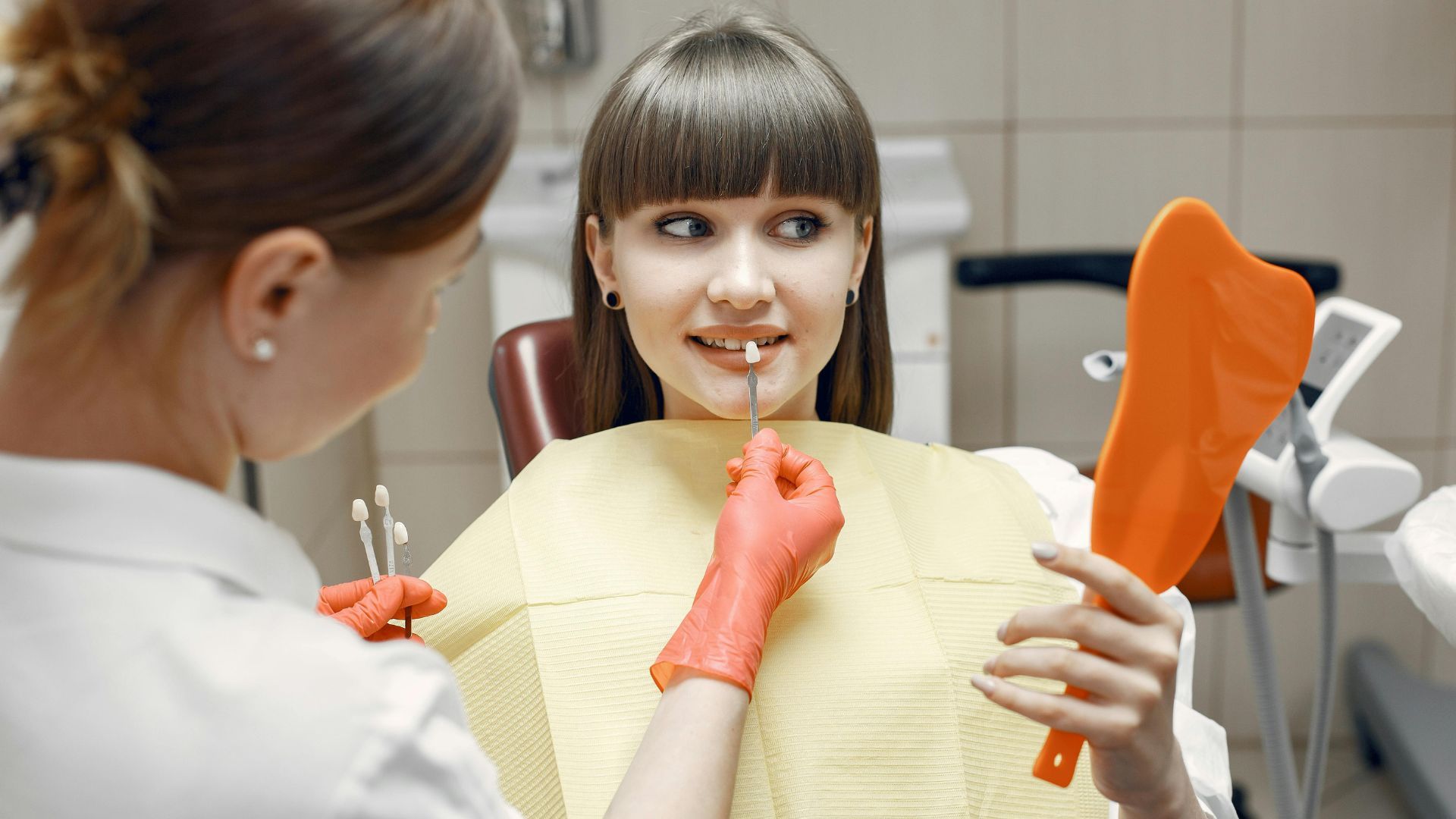 Dental hygienist in blue scrubs smiling beside a patient in a dental chair, with clinic tools in the background