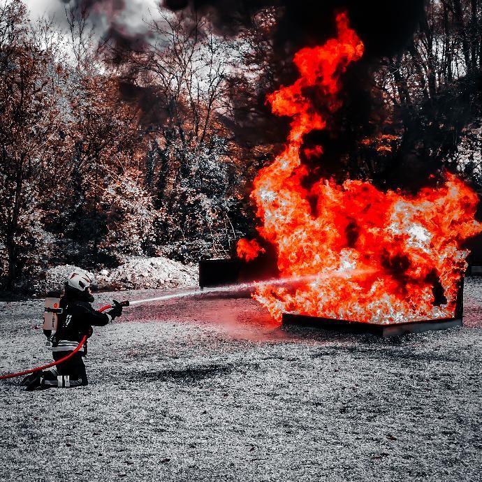 Een brandweerman knielt en spuit water op een groot, brandend plein in een openluchtomgeving.