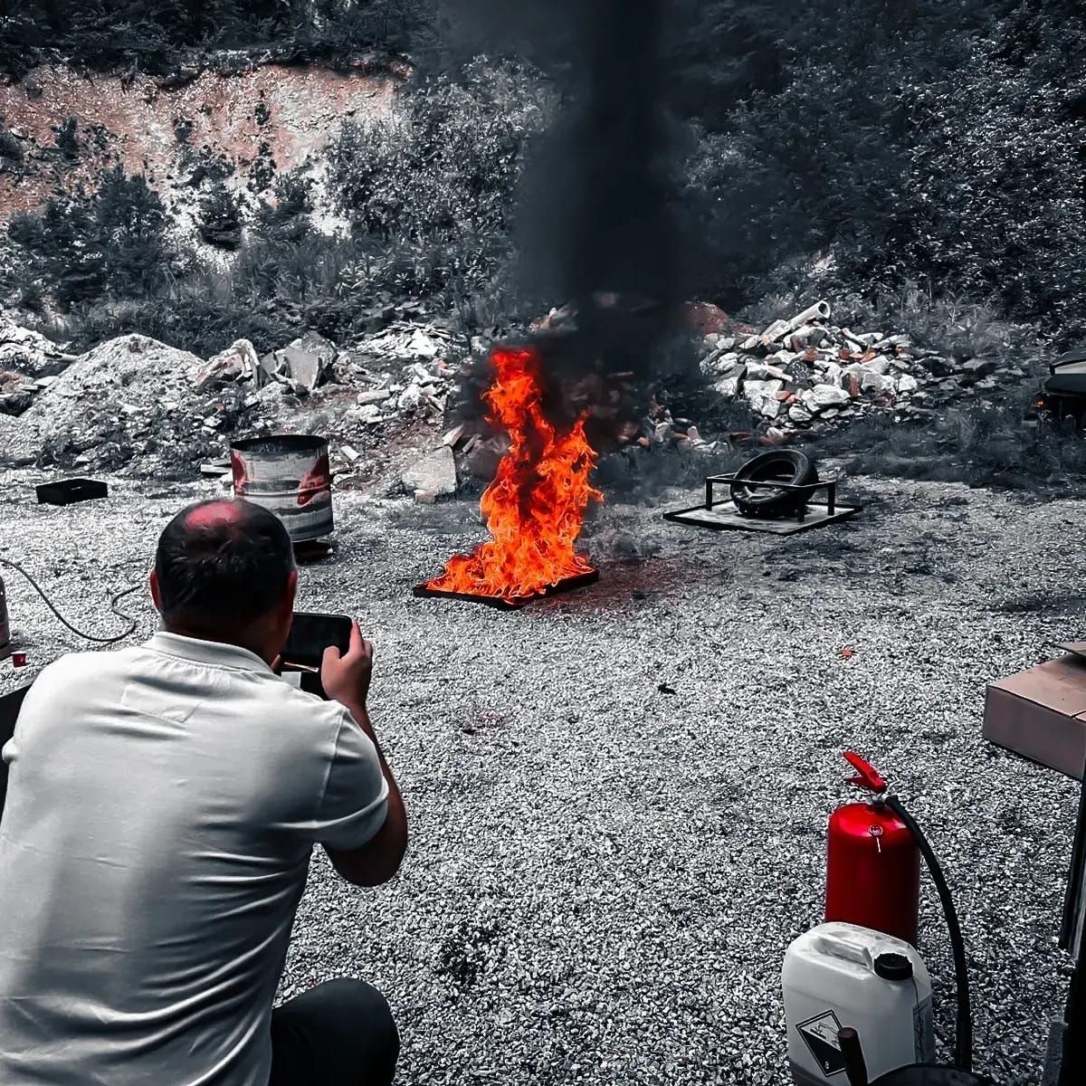 Een man fotografeert een brandend voorwerp in de buitenlucht; een brandblusser en een container staan ​​in de buurt.
