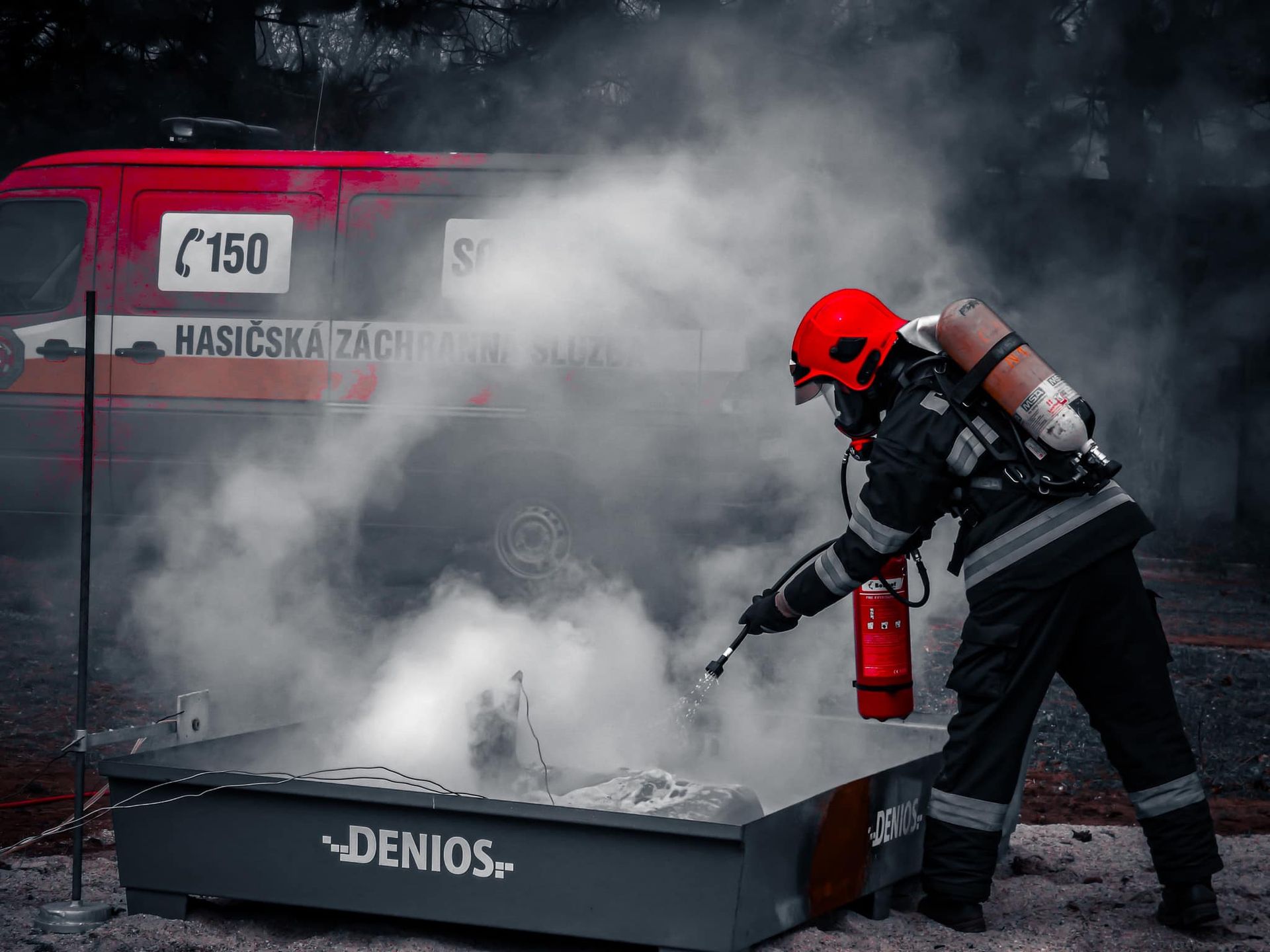 Brandweerman blust brand met een brandblusser, met rook en brandweerwagen op de achtergrond.