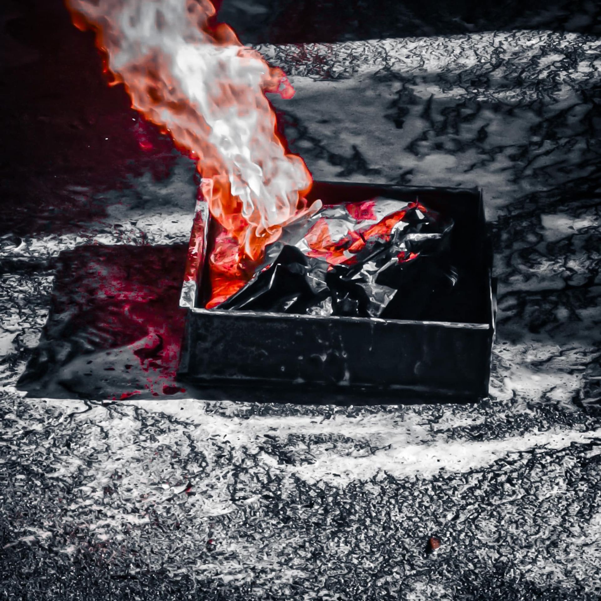 Een zwarte metalen container, gehuld in rode en witte vlammen, geplaatst tegen een zwart-witte rotsachtige ondergrond.