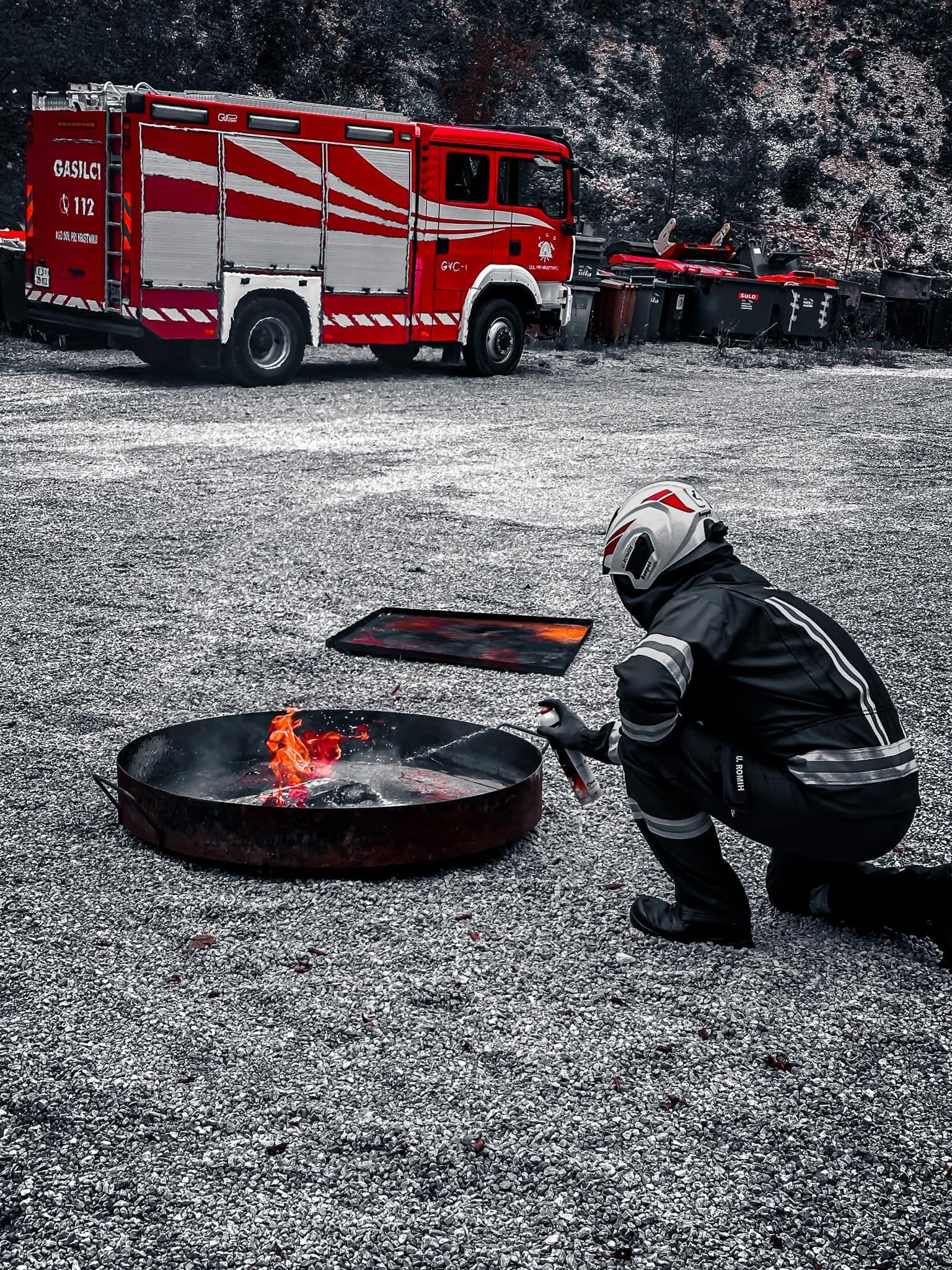 Knielende brandweerman steekt vuur aan in metalen vuurkorf, met brandweerwagen op de achtergrond.