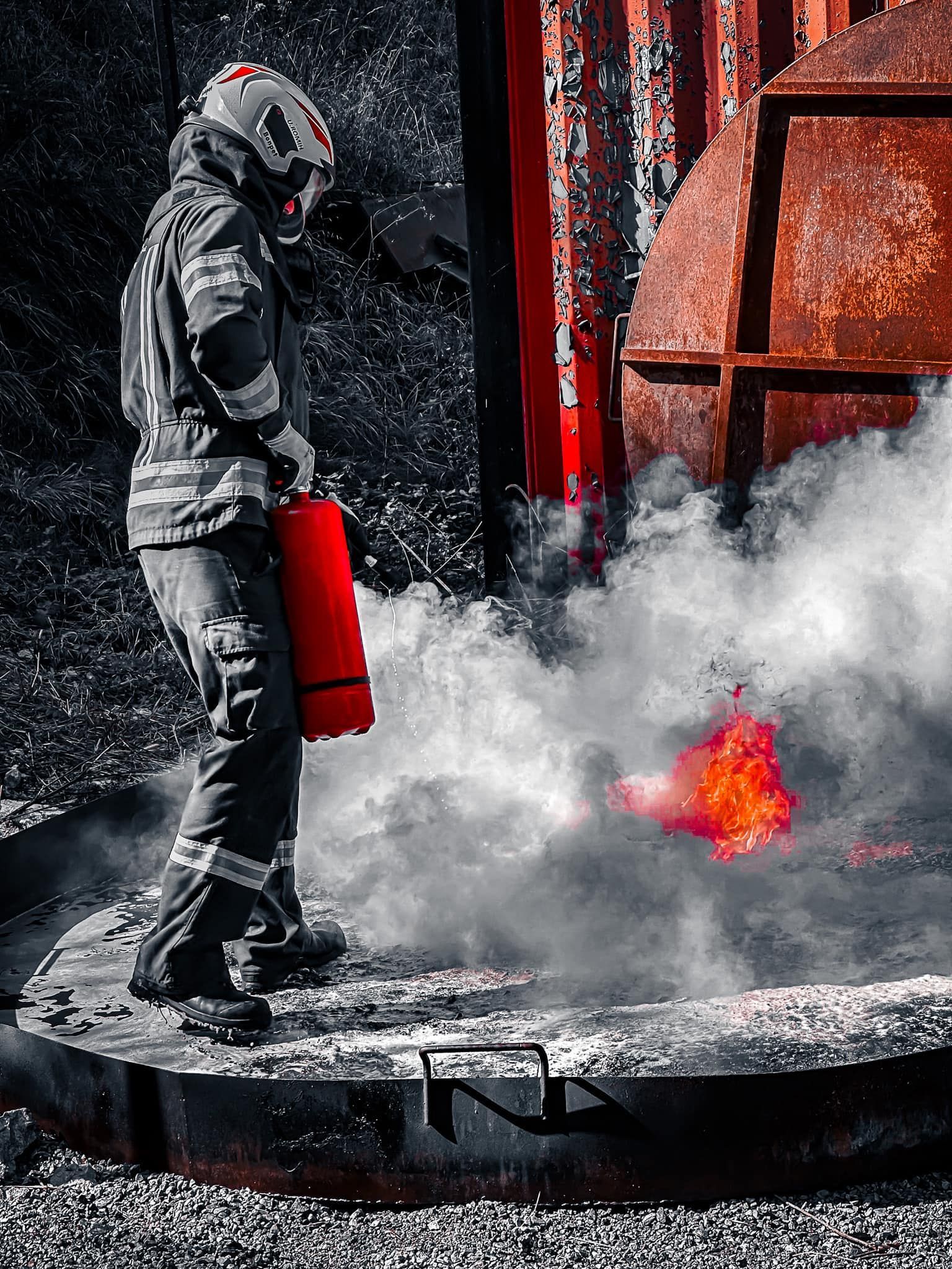 Een brandweerman in beschermende kleding gebruikt een rode brandblusser op een brandend voorwerp, omgeven door rook.