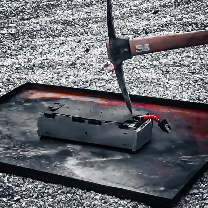 Een houweel slaat op een grijs accupakket op een roodbevlekte zwarte ondergrond, omgeven door grind.