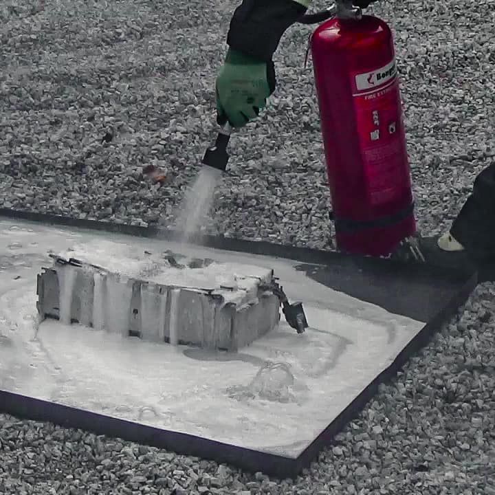 Iemand blust een metalen voorwerp met een brandblusser; wit schuim bedekt het voorwerp en de mat.