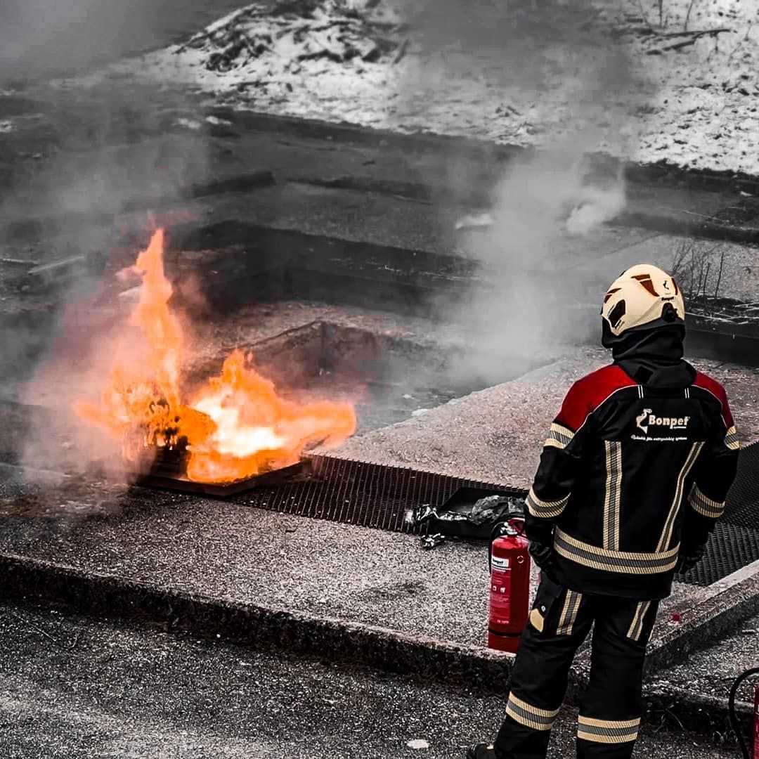 Een brandweerman in uniform ziet vlammen uit een rooster komen, met een brandblusser in de buurt en rook.