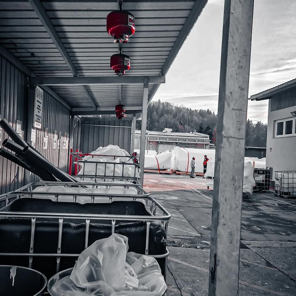 Industrieterrein in de openlucht met afgedekte containers, rode verlichting en arbeiders bij grote witte zakken.