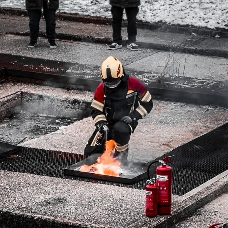 Een brandweerman knielt en blust een klein brandje met een brandblusser. Buiten.