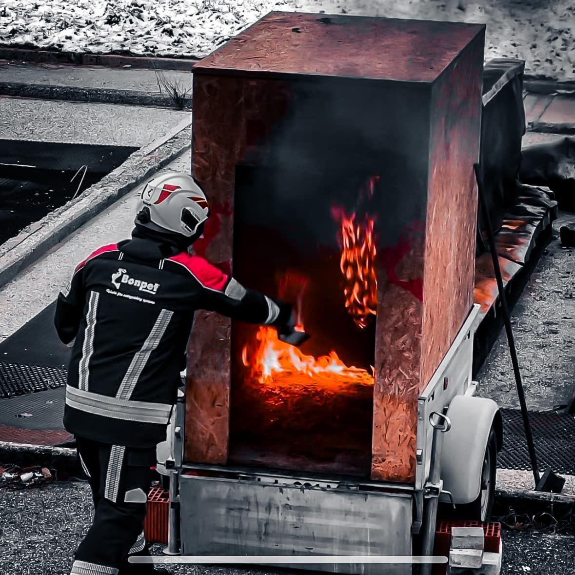 Brandweerman in rood-zwarte uitrusting bestrijdt brand in een houten kist op een aanhanger. Vlammen zijn zichtbaar.