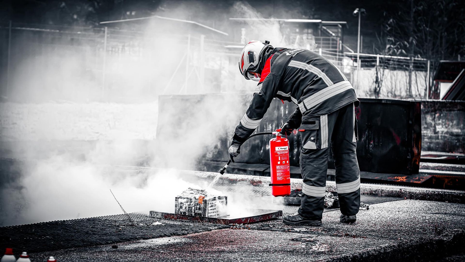 Een brandweerman in beschermende kleding blust een kleine brand met een rode brandblusser, omgeven door rook.