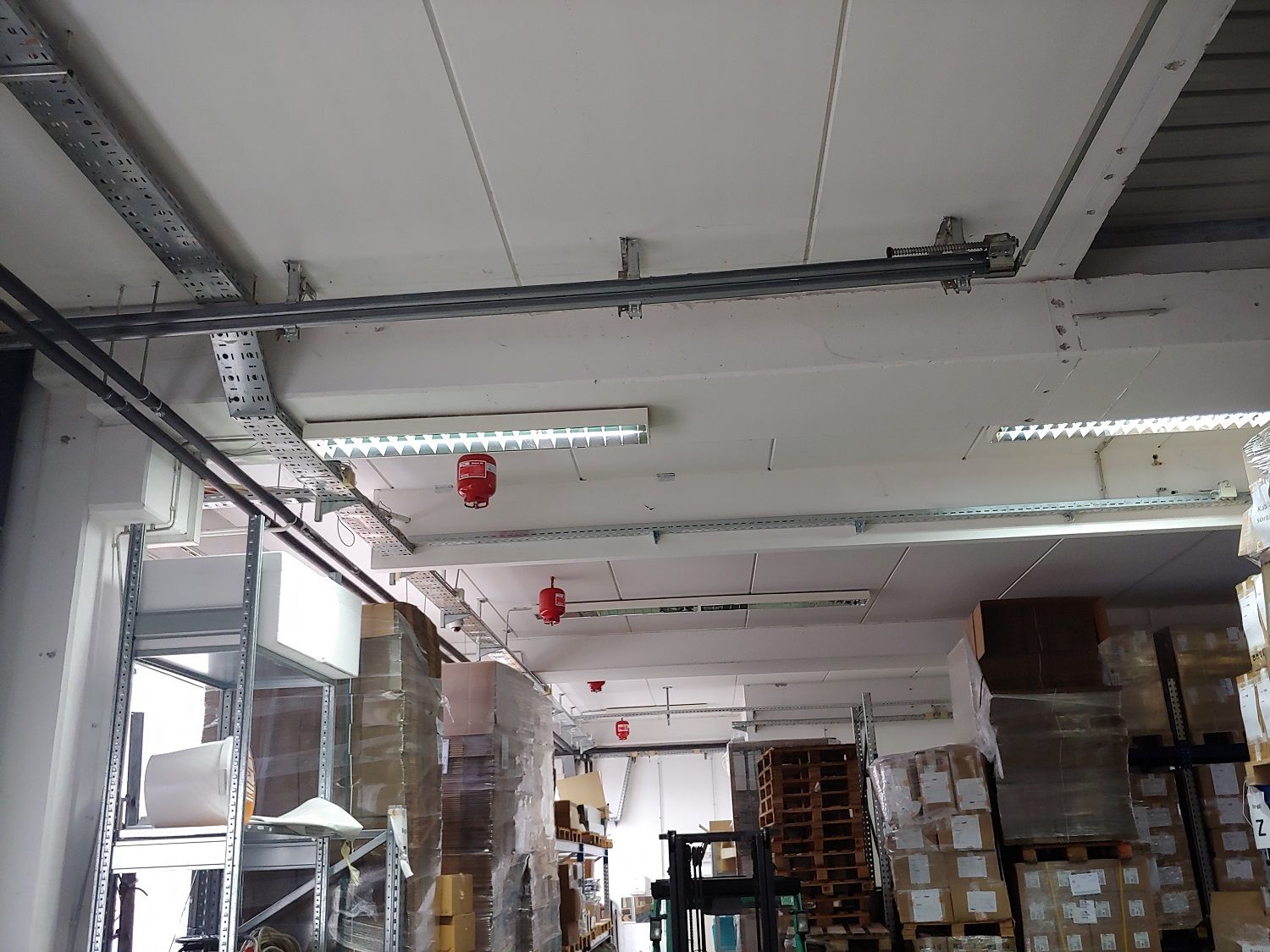 Magazijnplafond met elektrische leidingen, tl-lampen en rode brandmelders.