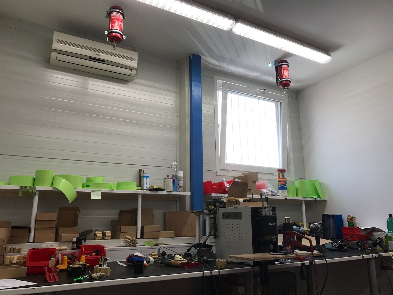 Werkplaatsinterieur met een rommelige werkbank, planken, airconditioningunit en hanglampen.