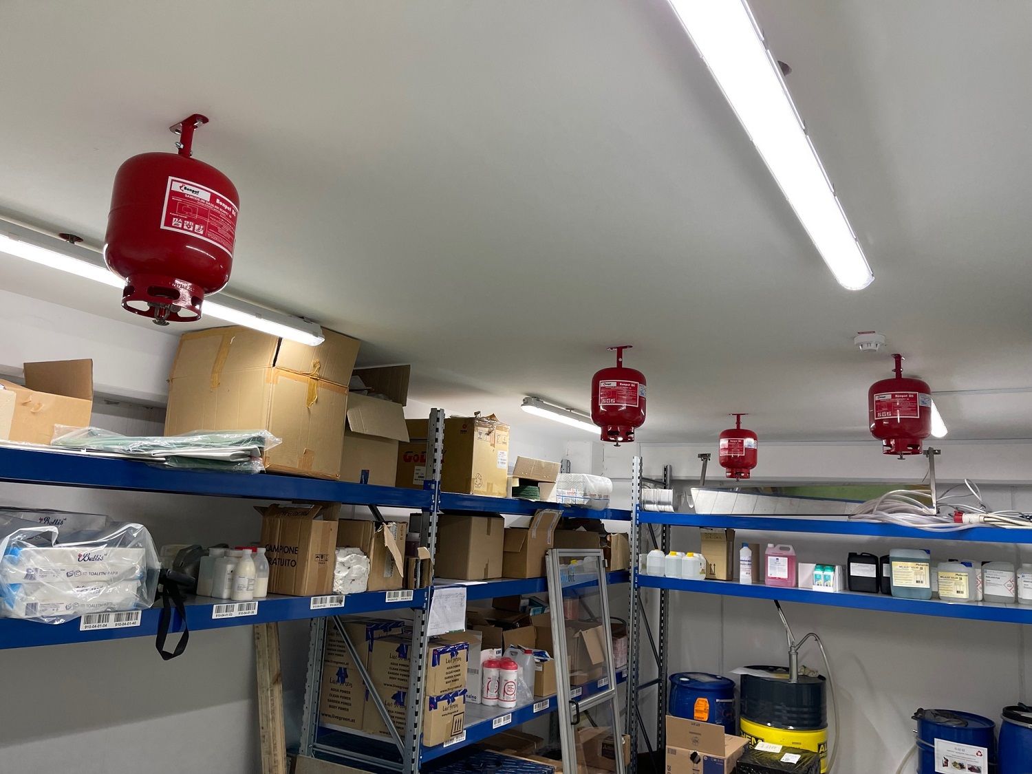 In een opslagruimte met schappen vol voorraden en dozen hangen rode brandblussers aan het plafond.