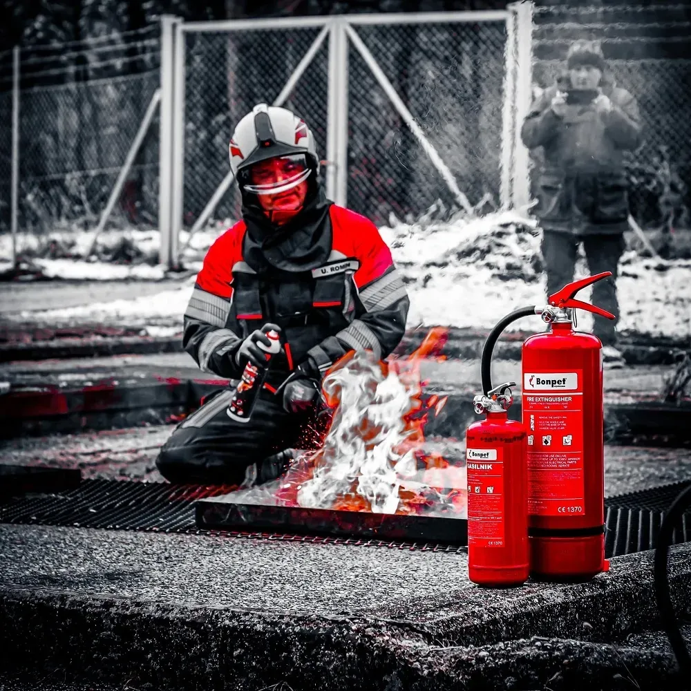 Brandweerman knielt en blust brand tijdens training, in rood pak en helm.