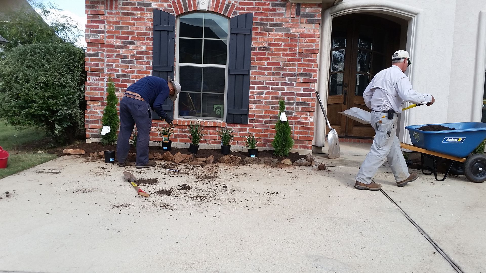 Two workers cleaning debris beside a brick house with a wheelbarrow and tools