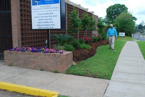 Sidewalk beside brick building with flower beds, a sign, and a person walking on the lawn path.