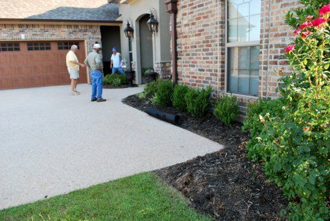 People standing near a brick house with a driveway and landscaped flower bed