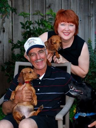 Smiling couple holding two small dogs and a black dog in a garden setting