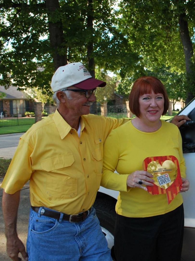 Two people standing outdoors by a car, both wearing bright yellow shirts, one holding a snack package.