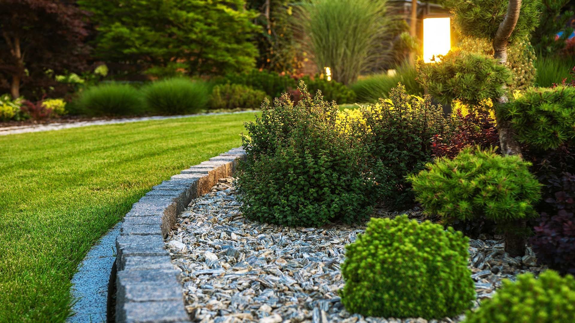 Curved garden bed with white stones, lush shrubs, and a glowing lantern beside a green lawn.