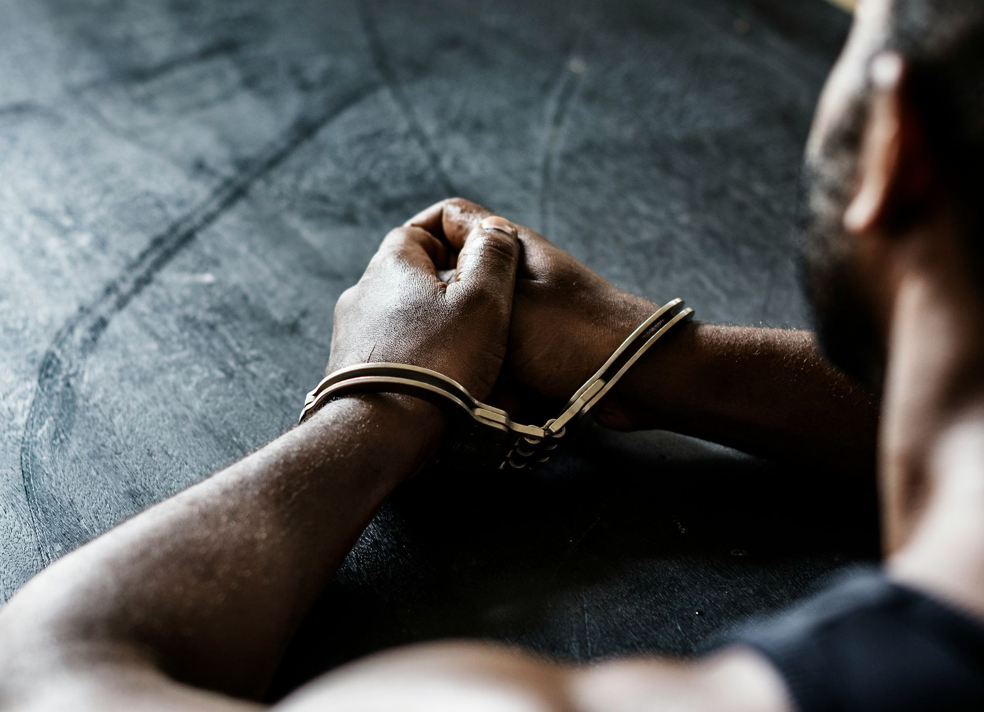 Person in handcuffs, hands clasped on a dark surface. Focus on the clasped hands and handcuffs.