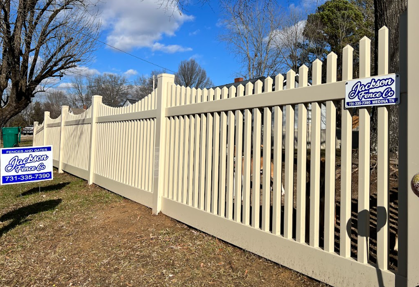 Cream-colored vinyl fence with pointed slats along a grassy roadside under a blue sky
