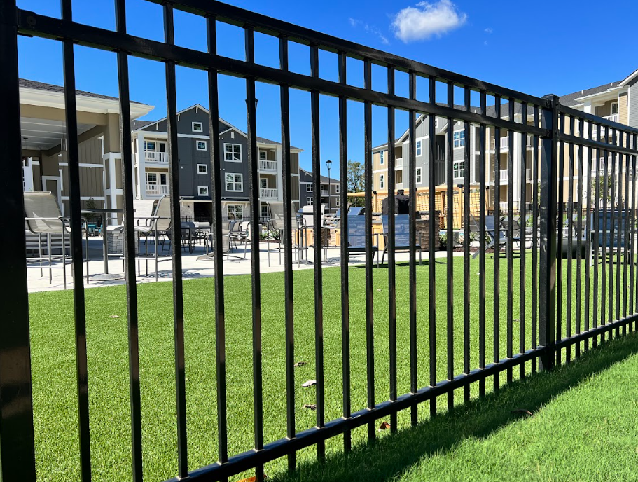 Black metal fence bordering a green lawn with apartment buildings and a pool in the background