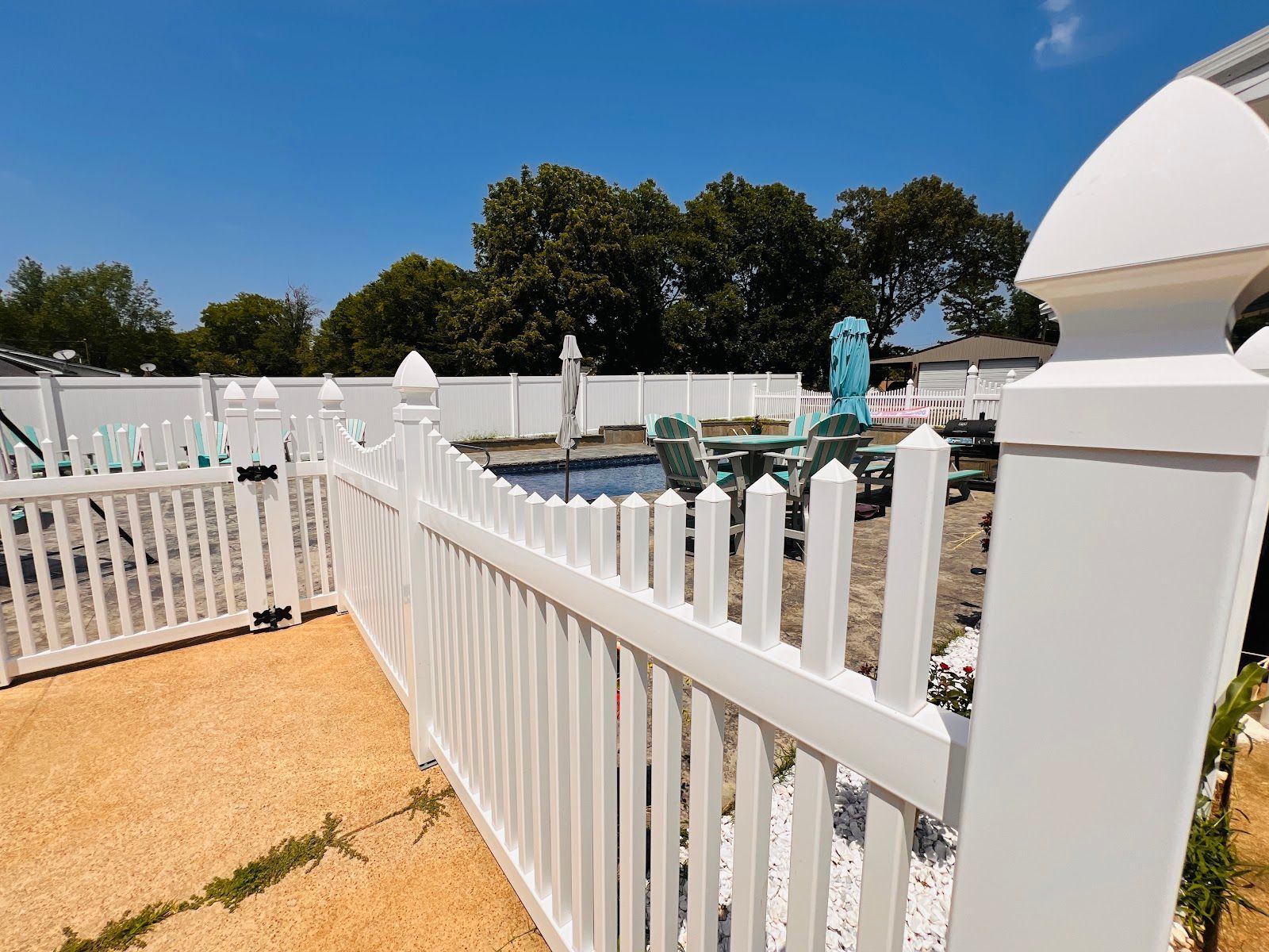 White fence beside a turquoise pool and white patio under a blue sky, with trees in the background