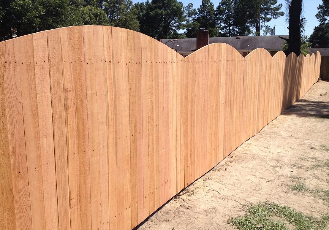 Wooden privacy fence with scalloped tops along a dirt path beside a yard.