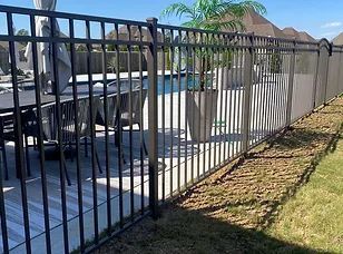 Black metal fence beside a patio and pool area, with grass and a sidewalk in sunlight