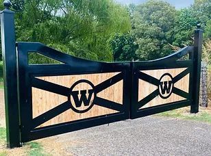 Black metal driveway gate with two large “W” emblems, set in a green outdoor area