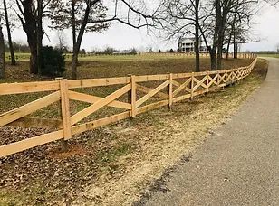 Wooden split-rail fence along a rural roadside with leafless trees and a distant farmhouse