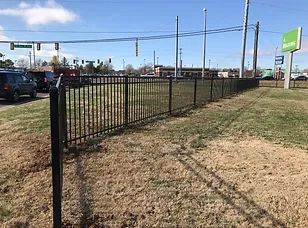 Black metal fence along a roadside lot under a clear sky, with cars and business signs in the background