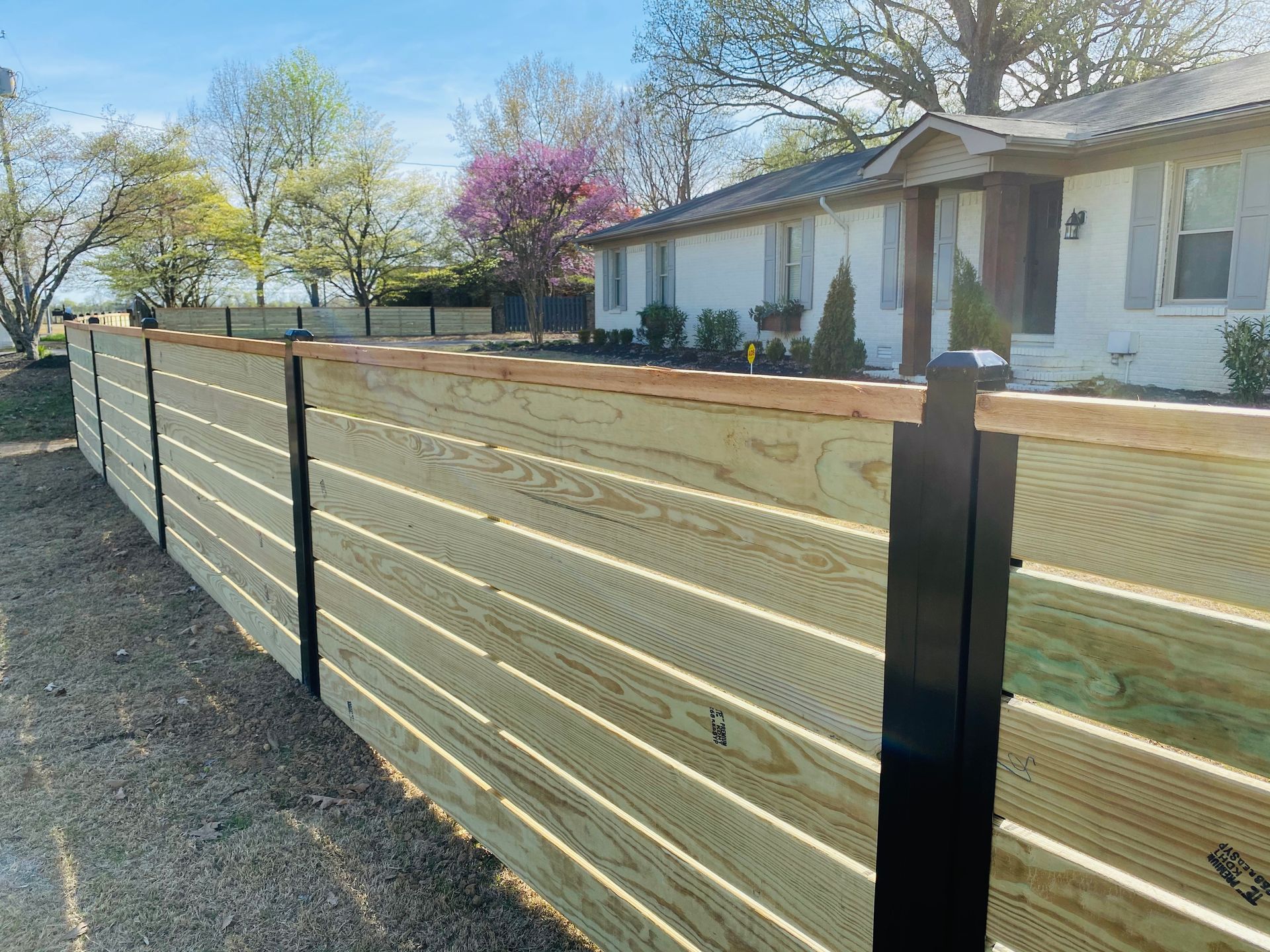 Wooden privacy fence beside a residential yard with a white house and blooming trees on a sunny day
